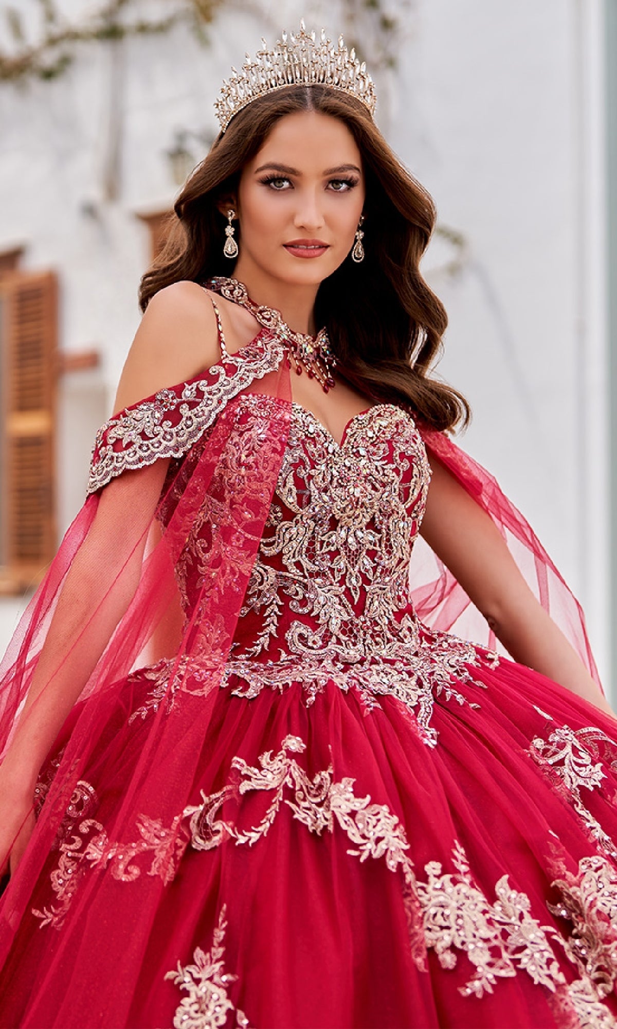 A woman in the Quinceanera Dress PR30086 By Princesa, featuring ornate silver embroidery and a jeweled crown, stands outdoors against a white building.